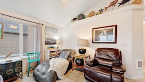 A living room with a leather sofa, a green chair, a small table with a lamp, a decorative shelf with items, and a window with blinds. - Modern Beach House with Partial Ocean Views - Walk to Nye Beach & Attractions (Newport)