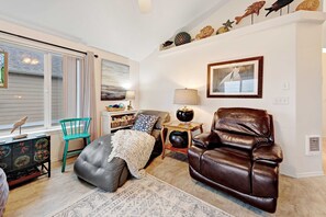 A living room with a leather sofa, a green chair, a small table with a lamp, a decorative shelf with items, and a window with blinds. - Modern Beach House with Partial Ocean Views - Walk to Nye Beach & Attractions (Newport)