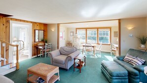 A spacious living room with a green carpet, wooden walls, a staircase, a dining area with a table and chairs, and a large window. - Beautiful, dog-friendly cottage with large yard - just a few blocks to the beach (Gearhart)