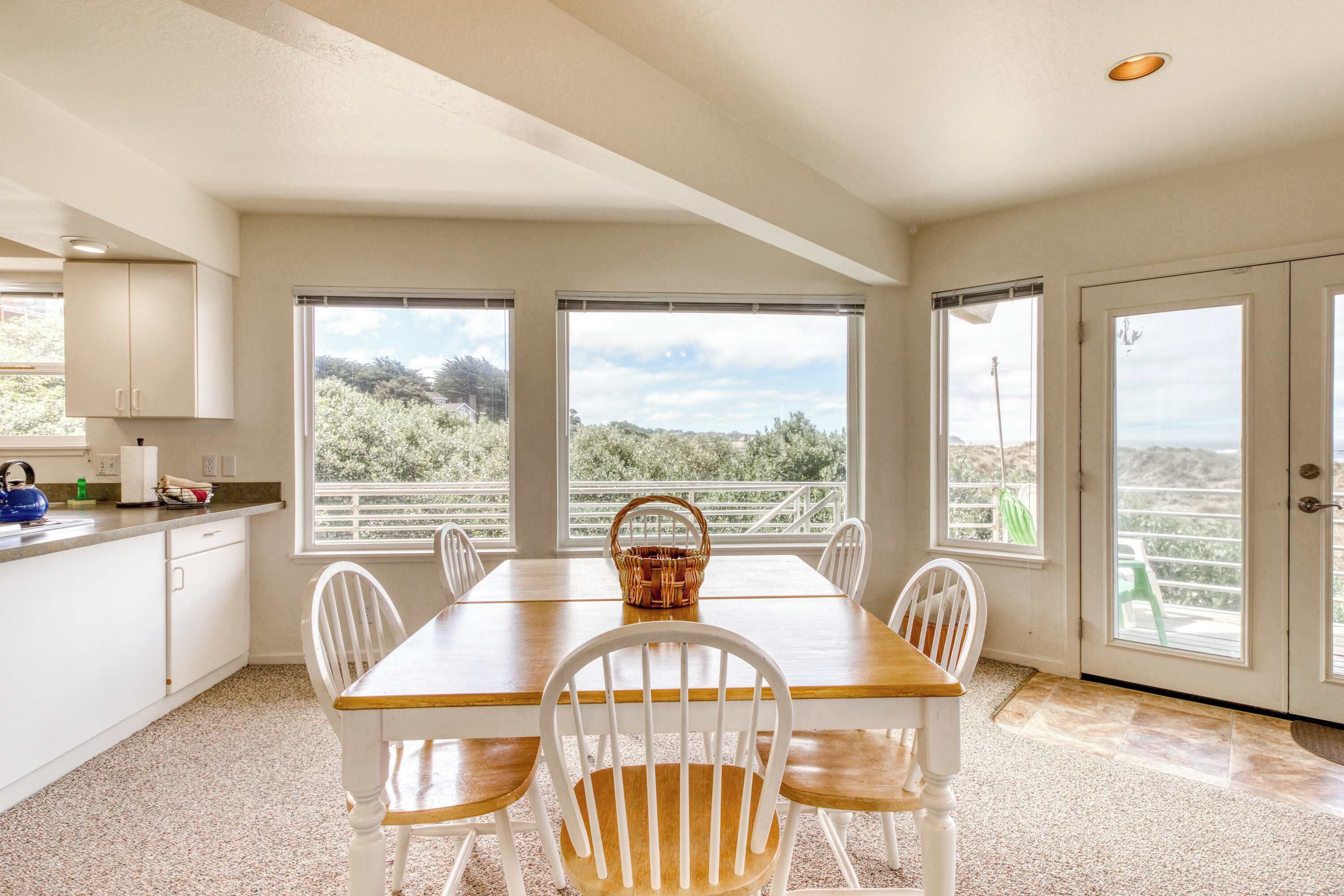 Une cuisine spacieuse avec une grande table à manger, des chaises blanches et une vue sur l’extérieur grâce à de grandes fenêtres.