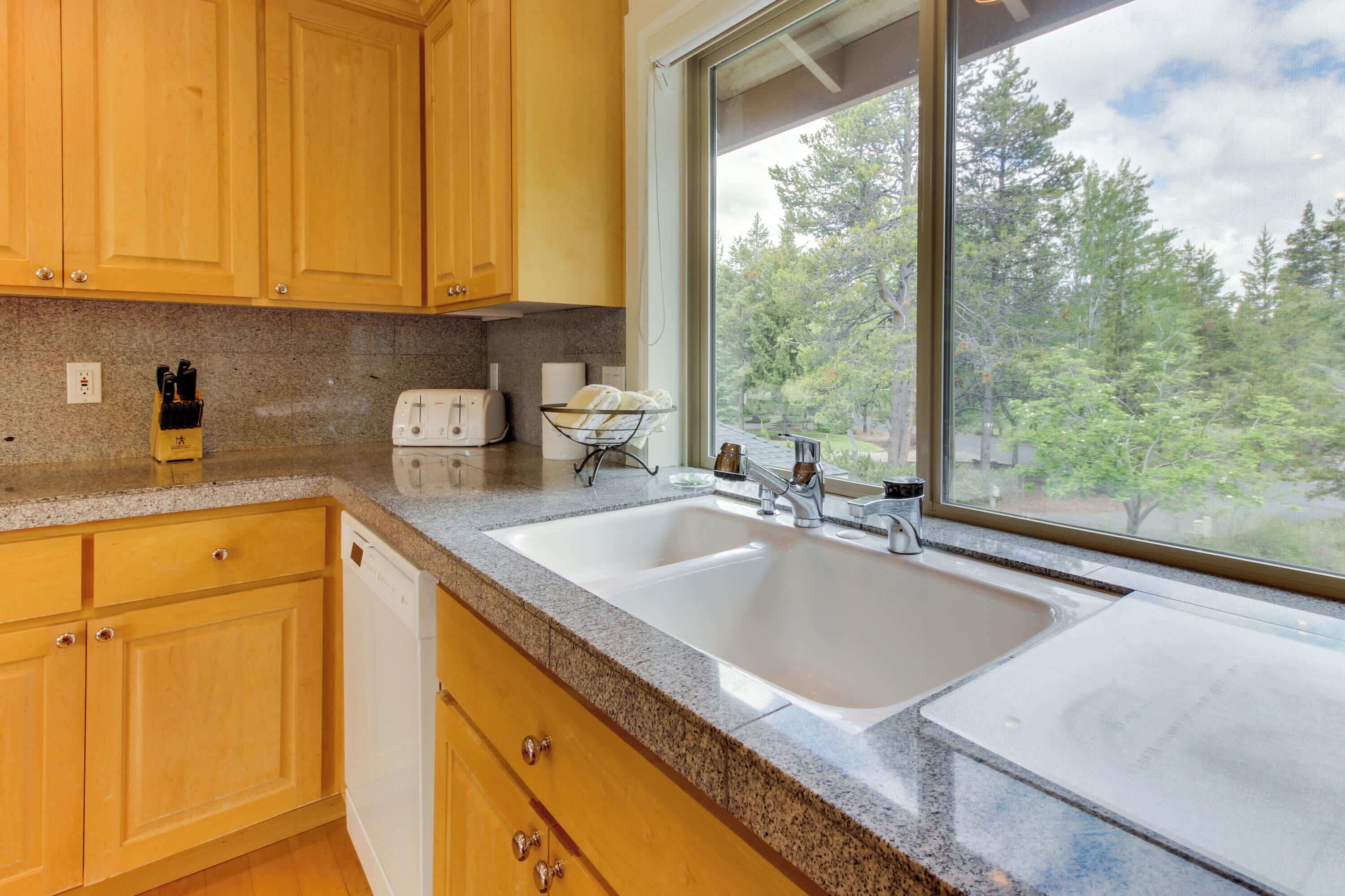 A kitchen with wooden cabinets, a granite countertop, a double sink, and a window with a view of trees.