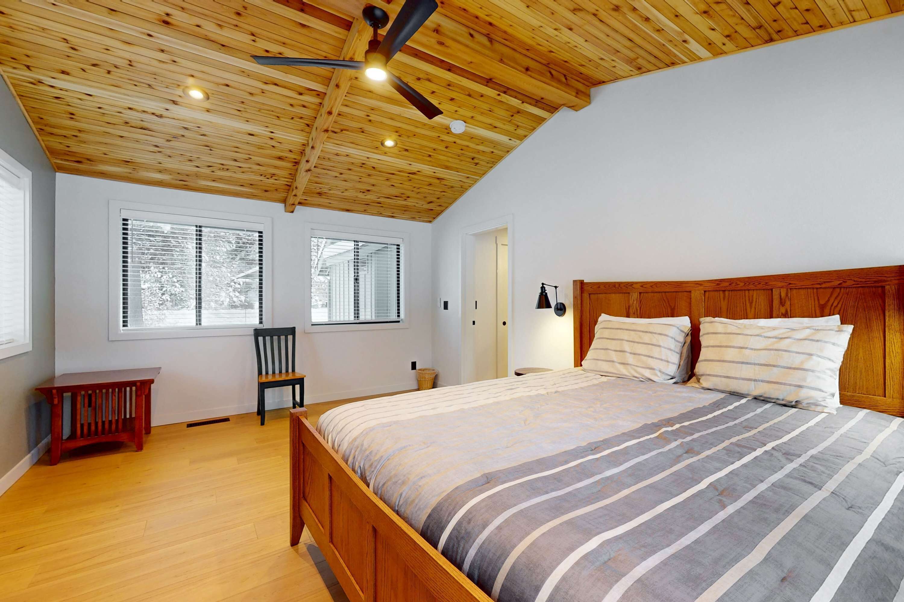 Une chambre avec un plafond en bois, un grand lit, deux fenêtres, une chaise et une petite table.
