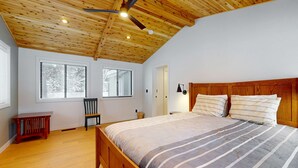 A bedroom with a wooden ceiling, a large bed, two windows, a chair, and a small table. - Comfy Home with Private Hot Tub, SHARC Passes & Entertainment (Sunriver)