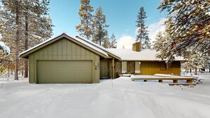 A two-story building with a garage, a covered porch, and a snow-covered landscape. - Comfy Home with Private Hot Tub, SHARC Passes & Entertainment (Sunriver)