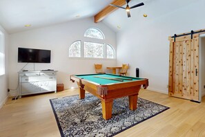 Une pièce équipée d’une table de billard, d’un téléviseur, d’une armoire en bois et d’une grande fenêtre donnant sur la forêt.