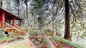 Una cabana de fusta amb façana vermella, una escala de fusta i un camí envoltat per un bosc dens i un riu.