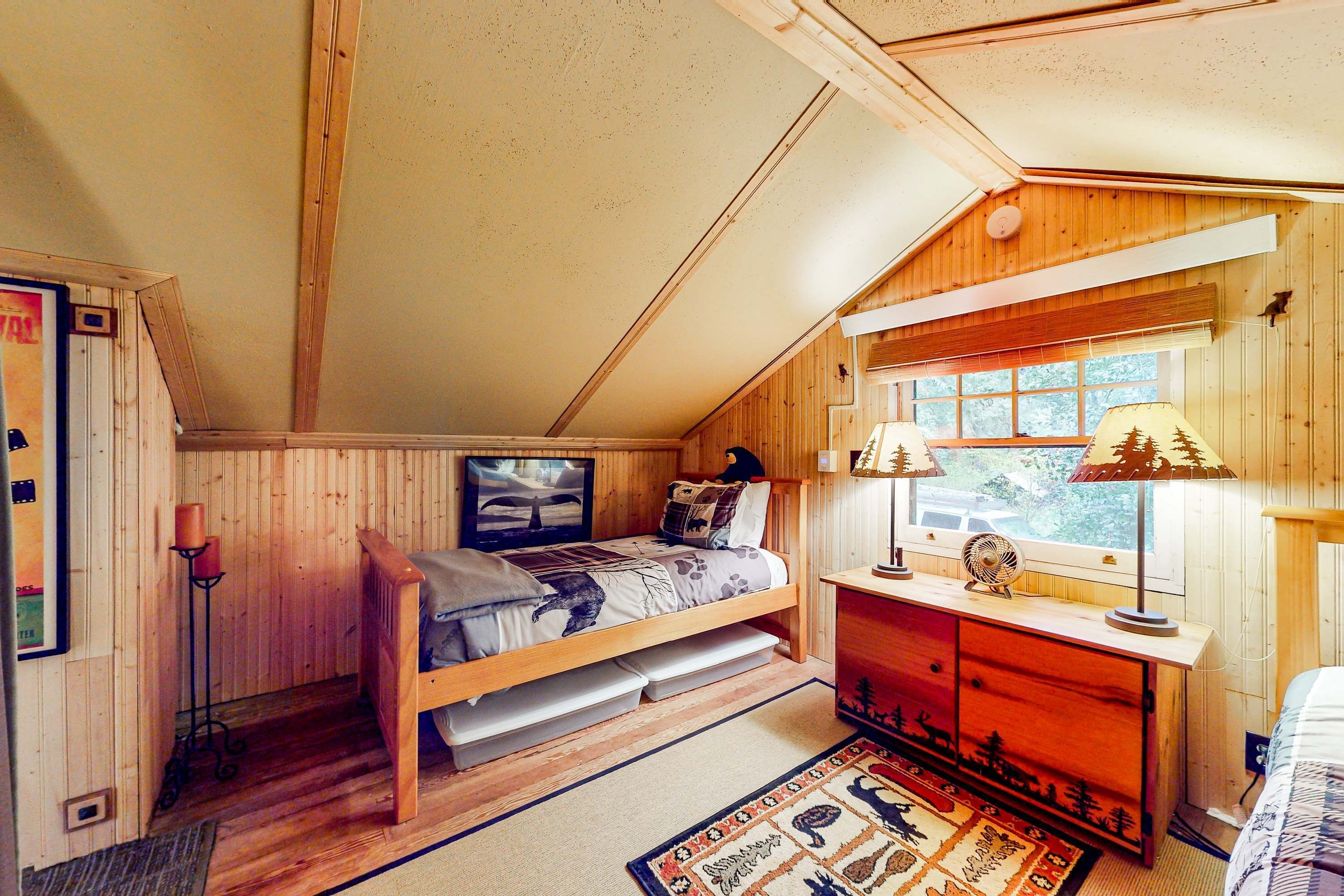 Ein gemütliches Schlafzimmer mit einem Holzbett mit Etagen, einem Schreibtisch mit Lampe und einem Fenster mit Blick auf Bäume.