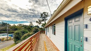 Eine Holzterrasse führt zu einem Gebäude mit einer grünen Tür, umgeben von Grünpflanzen und einem bewölkten Himmel.