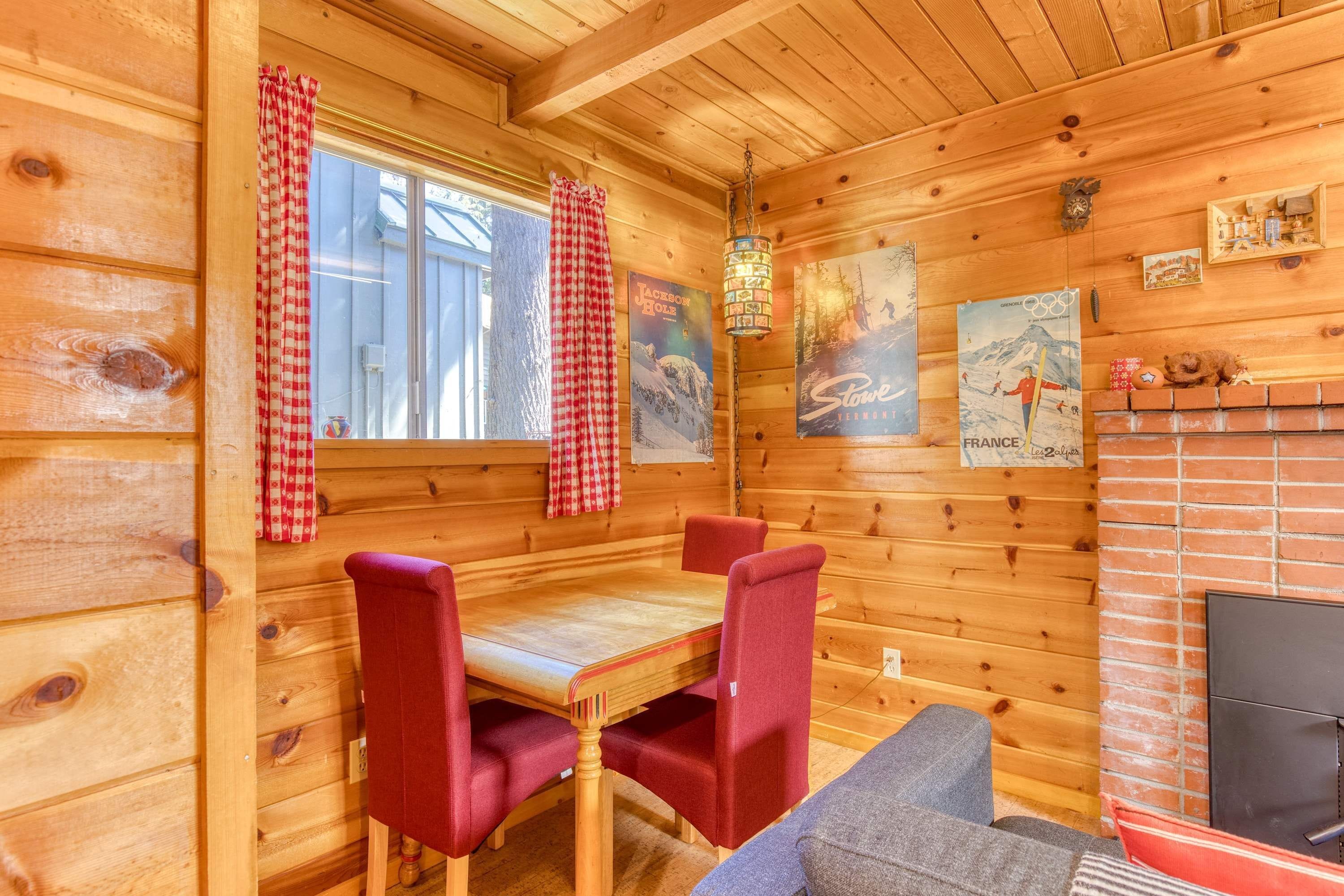 L’intérieur d’un chalet en bois comprend une table à manger, des chaises à revêtement rouge, une cheminée et des éléments de décoration murale, notamment une affiche représentant les anneaux olympiques et un paysage de montagne.