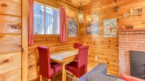 Ein Holzhütten-Interieur mit einem Esstisch, rot gepolsterten Stühlen, einem Kamin und Wanddekorationen, darunter ein Poster mit den Olympischen Ringen und einer Berglandschaft.