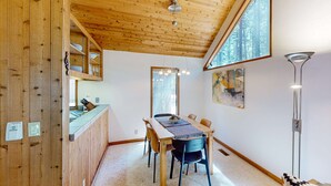 Un plafond en bois, une table à manger avec des chaises, un luminaire suspendu et une fenêtre donnant sur des arbres.
