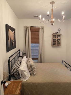 A bedroom with a bed, a window with blinds, a chandelier, a wall-mounted cabinet with figurines, and a painting of a cow.
