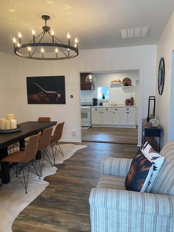 A modern living room with a dining table, sofas, a chandelier, and a kitchen in the background.