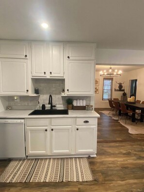 A modern kitchen with white cabinetry, a built-in dishwasher, and a dining area with a wooden table and chairs.