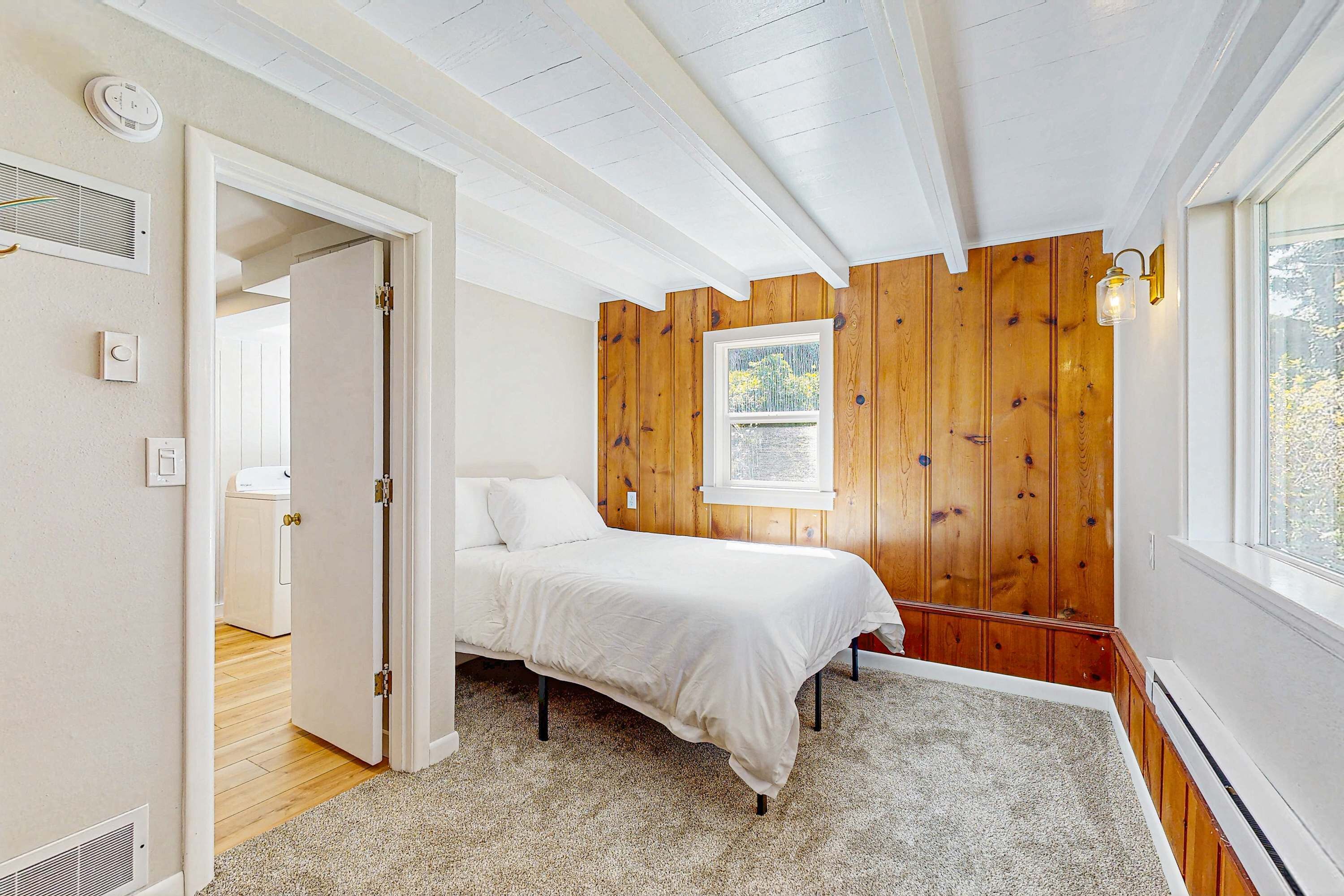 A bedroom with a bed, a window, wooden paneling, and a door leading to another room.