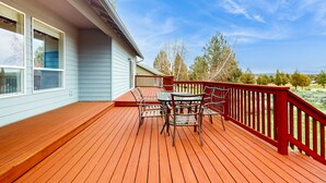 Eine Holzterrasse mit Tisch und Stühlen, einem roten Geländer sowie Blick auf Bäume und einen klaren Himmel.