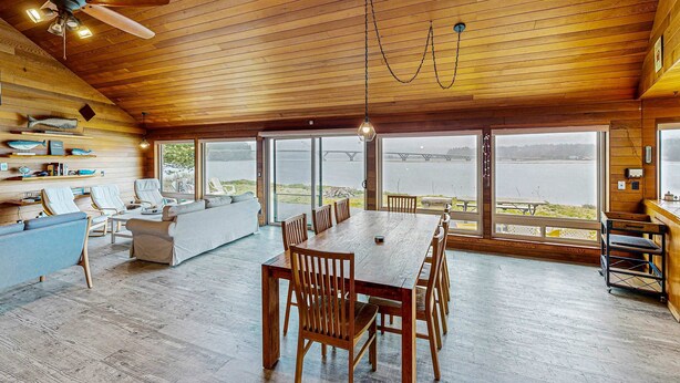 Une pièce spacieuse avec un plafond en bois, de grandes fenêtres, une table à manger avec des chaises, un canapé et un ventilateur de plafond.
