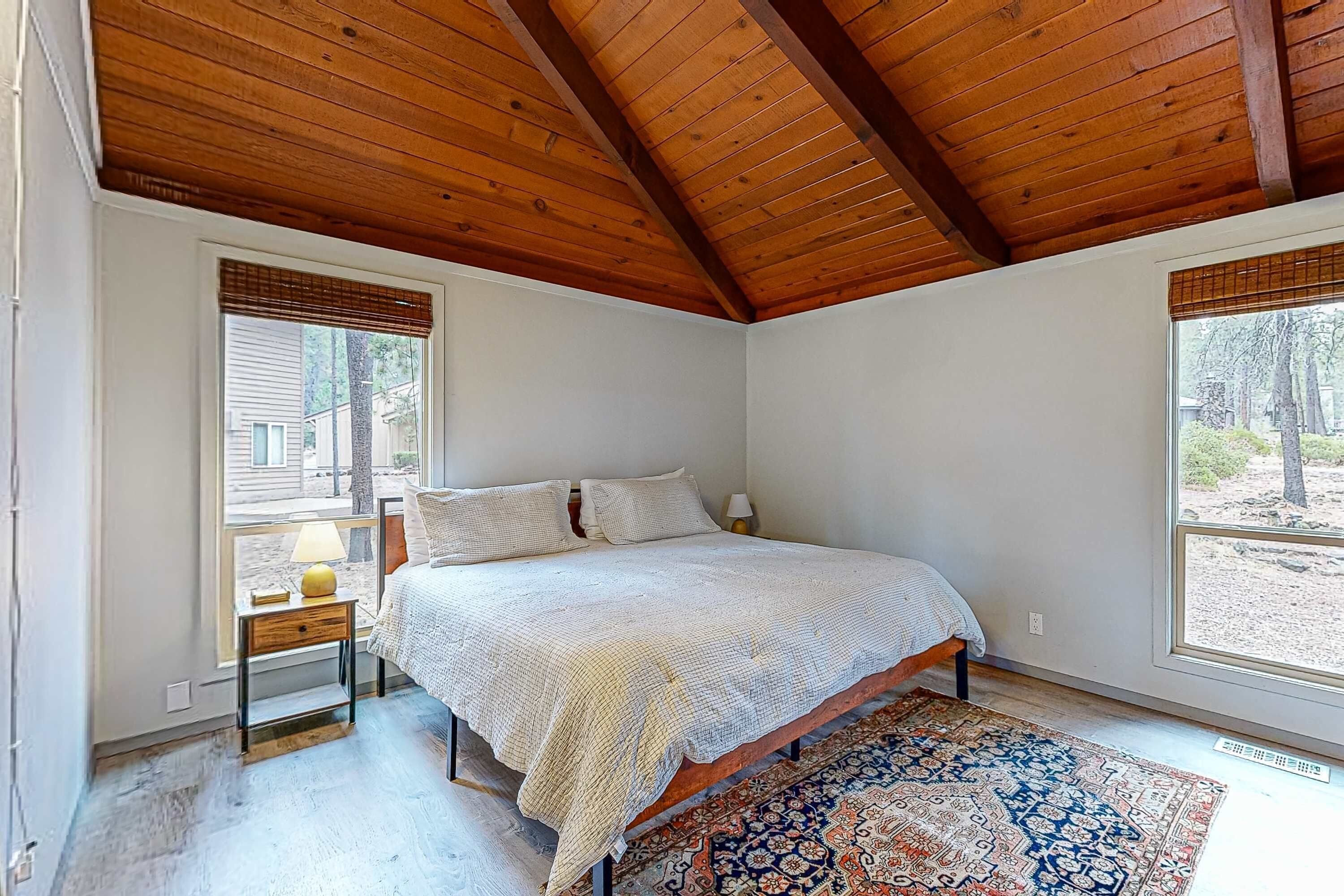 Una camera da letto con soffitto in legno, un letto con copripiumino fantasia, un comodino con una lampada e un'ampia finestra vista alberi.