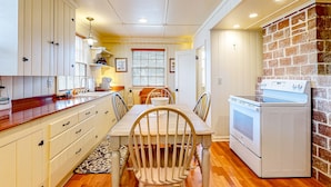 A compact kitchen with wooden cabinets, a dining table, chairs, a washing machine, and a brick wall. - Beautiful Cape Cod cottage - one block from the beach with covered balcony & W/D (Seaside)