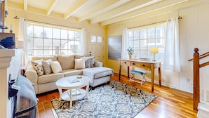 A spacious living room with a high ceiling, a sofa, a coffee table, a desk, and a staircase. - Beautiful Cape Cod cottage - one block from the beach with covered balcony & W/D (Seaside)