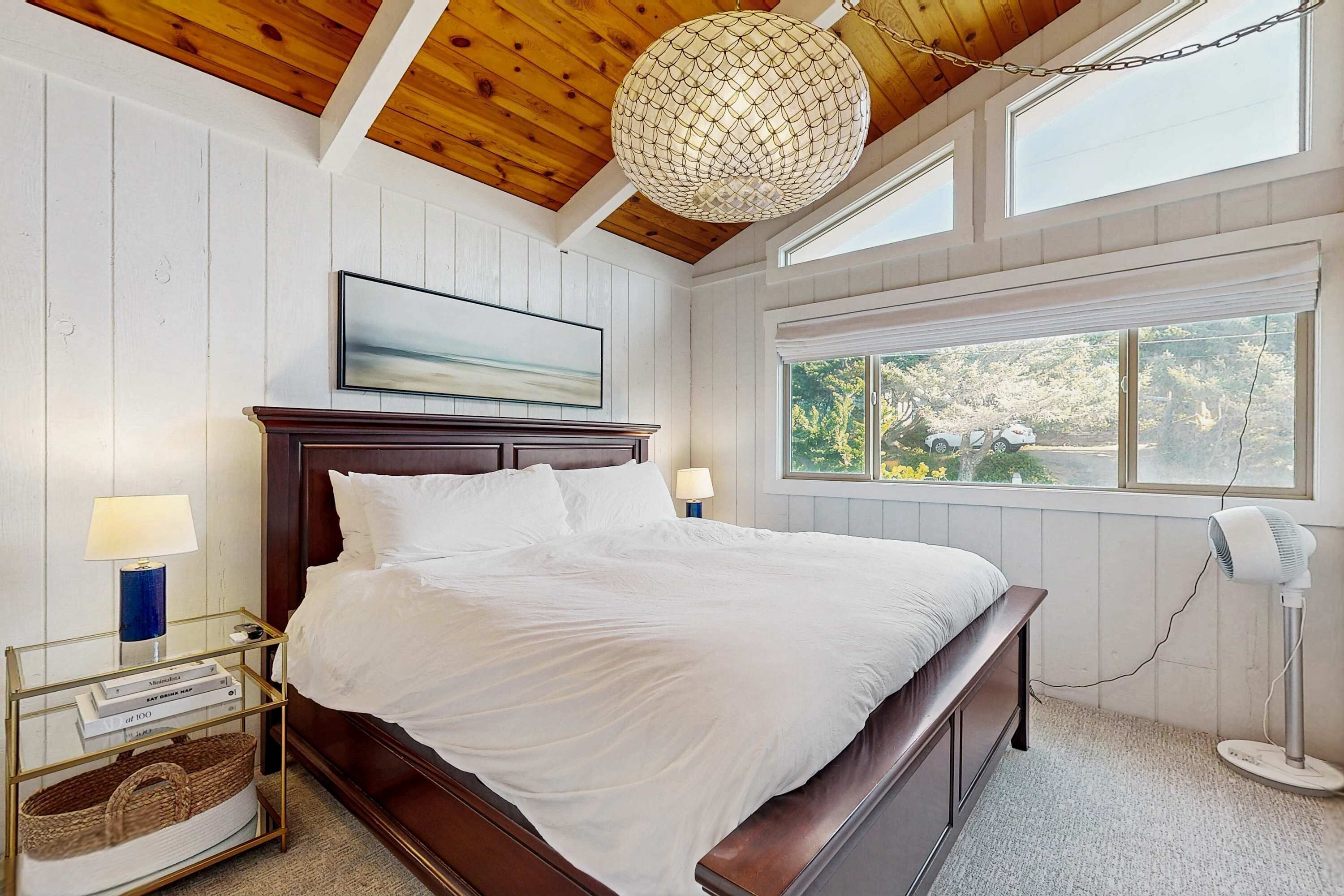 Quarto com teto de madeira, cama com roupa de cama branca, mesa de cabeceira com abajur, quadro na parede e janela com vista para árvores.
