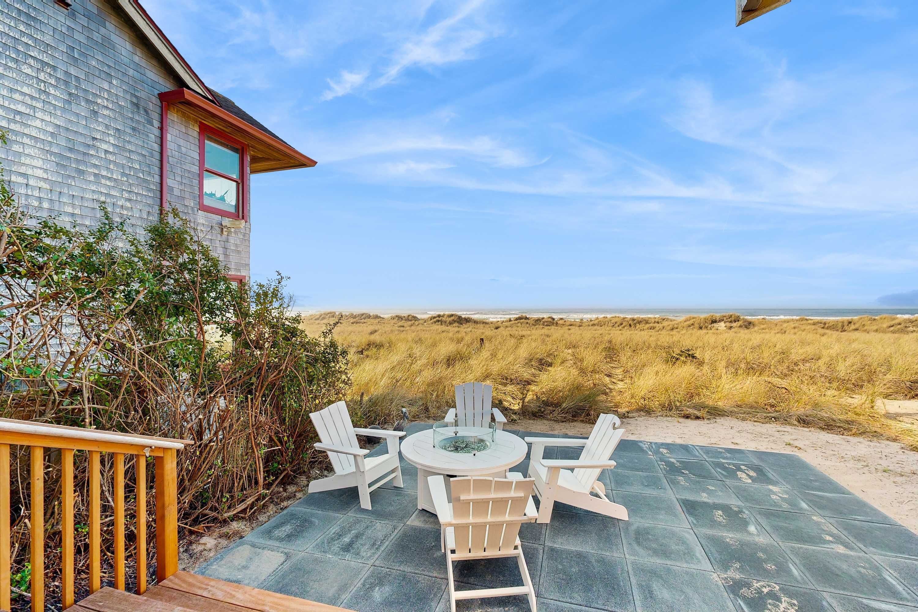 A patio with a round table and four Adirondack chairs, a wooden deck, and a view of the beach and dunes.