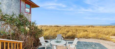 A patio with a round table and four Adirondack chairs, a wooden deck, and a view of the beach and dunes.