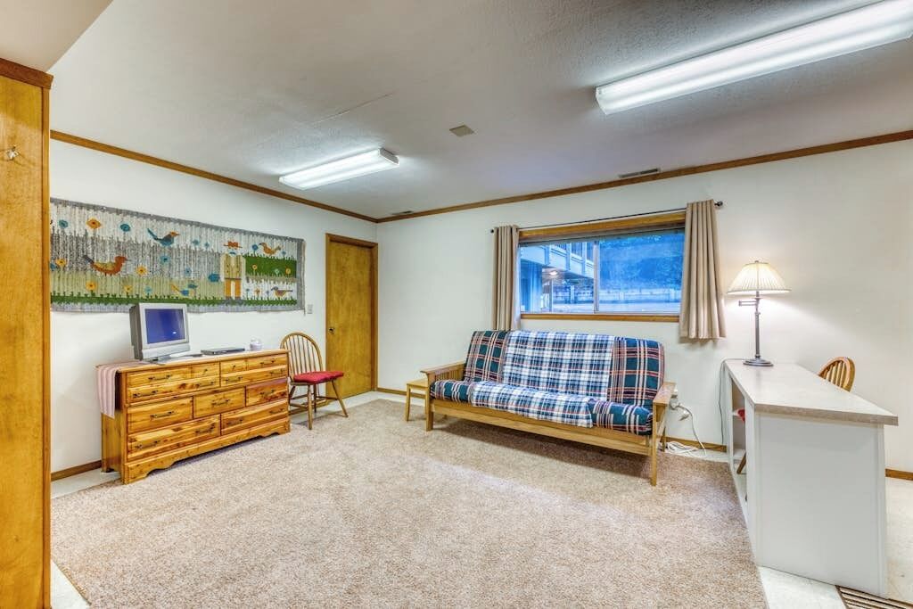 A room with a wooden dresser, a plaid sofa, a television, a desk, and a window with a view.