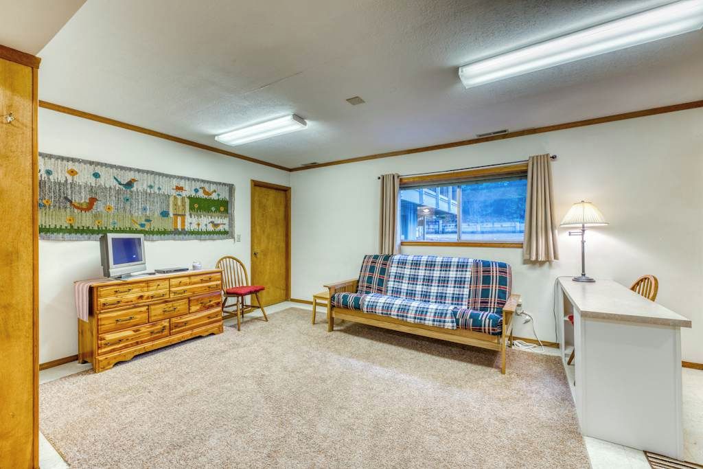 A room with a wooden dresser, a plaid sofa, a television on a stand, a window with curtains, and a lamp.