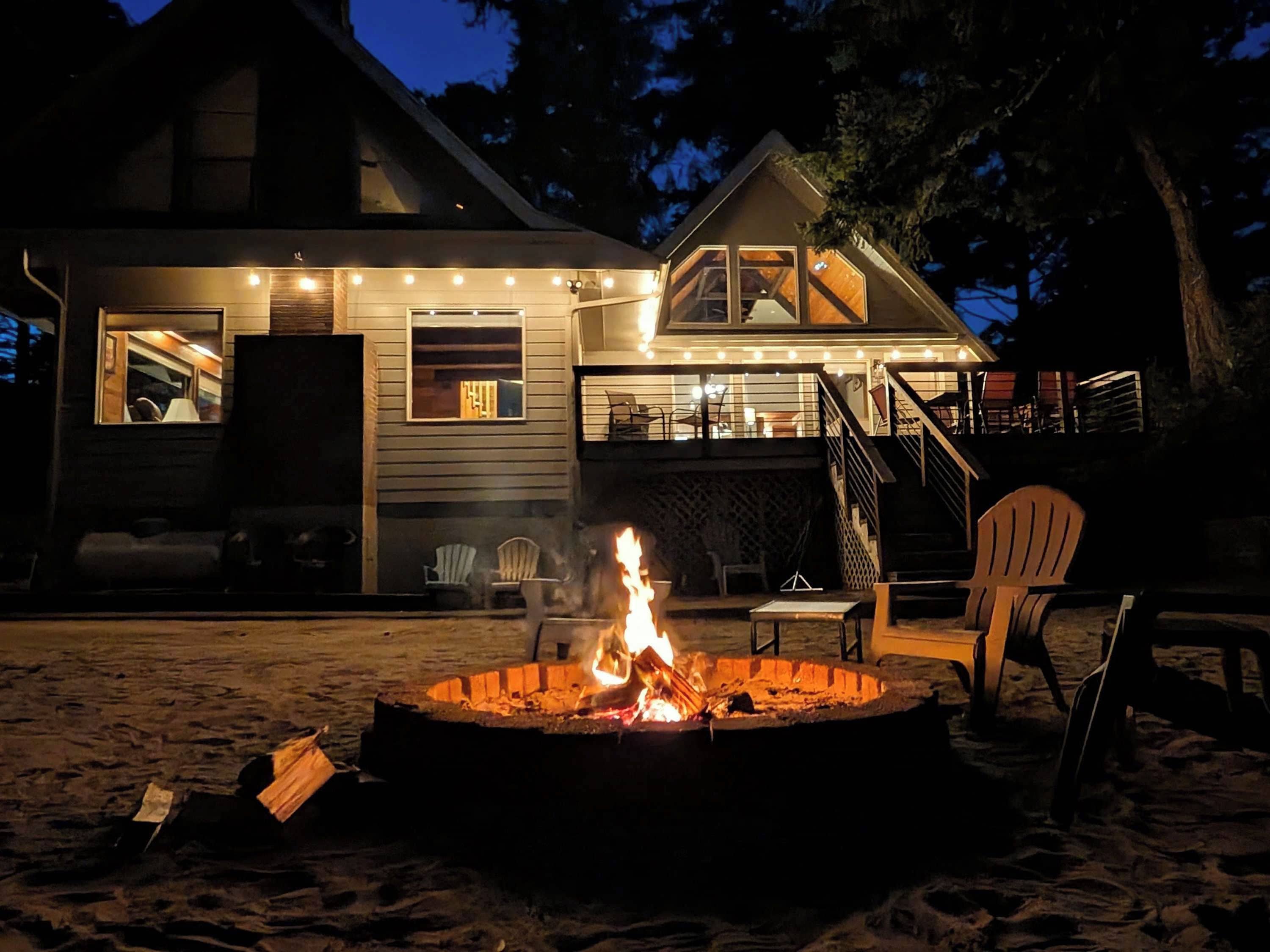 A two-story house with a fire pit in the foreground, outdoor seating, and a deck with string lights.