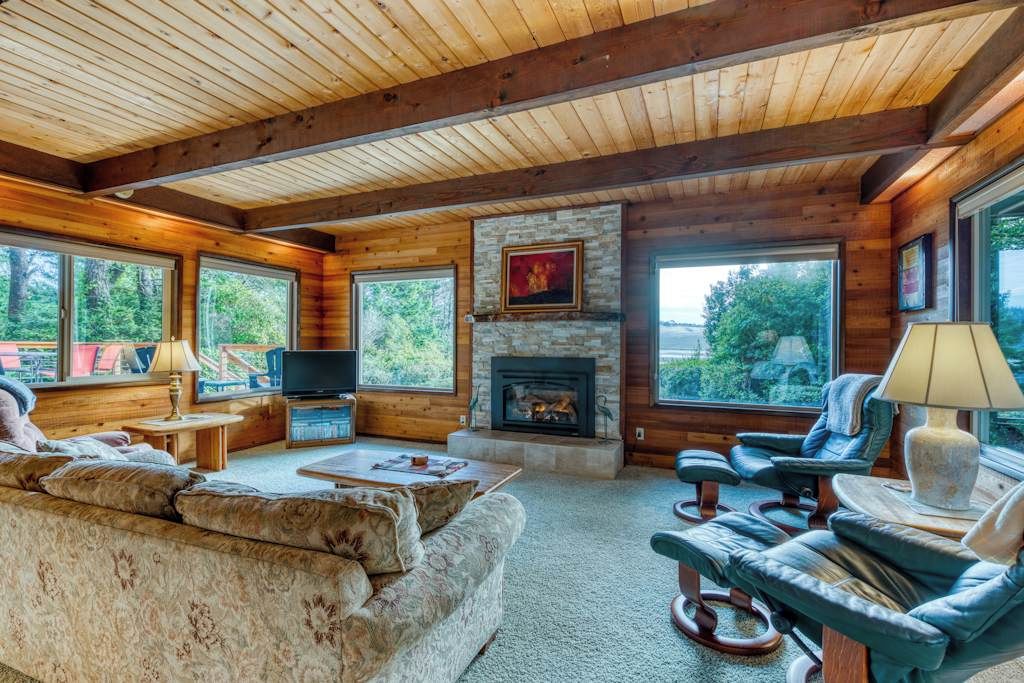 A log cabin interior with a fireplace, large windows, a sofa, and a television.