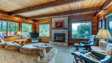 A log cabin interior with a fireplace, large windows, a sofa, and a television.