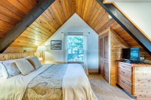A bedroom with a sloped ceiling, a bed with patterned bedding, a dresser with a television, and a window with a view of trees. - Gorgeous riverfront house with entertainment - near Bay Street and the beach (Florence)