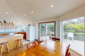 Una cocina moderna con encimeras de madera, una isla central con taburetes, una zona de comedor con mesa y sillas de madera, y un balcón con vistas a zonas verdes.