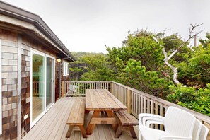 Una terraza de madera con mesa y sillas de picnic, rodeada por una barandilla de madera y abundante vegetación.