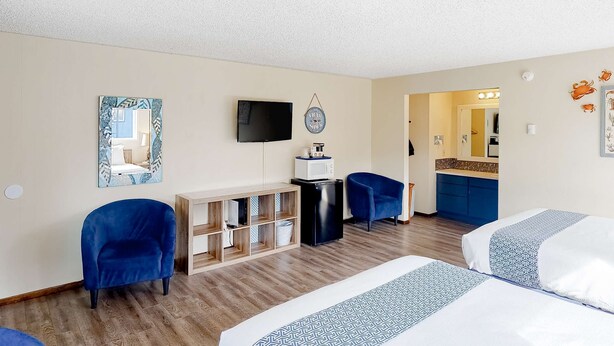 Ein Hotelzimmer mit zwei Betten, einem Fernseher, einem Schreibtisch mit einer Mikrowelle, einem Sessel und einer kleinen Küchenzeile.