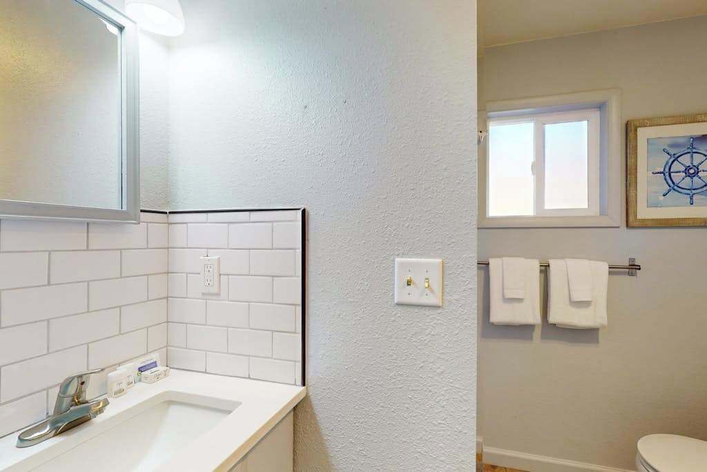 Ein Badezimmer mit einem weißen Waschbecken, einem Spiegel und weißen Fliesenwänden. Ein Fenster bietet Blick ins Freie, und ein gerahmtes Bild hängt an der Wand.