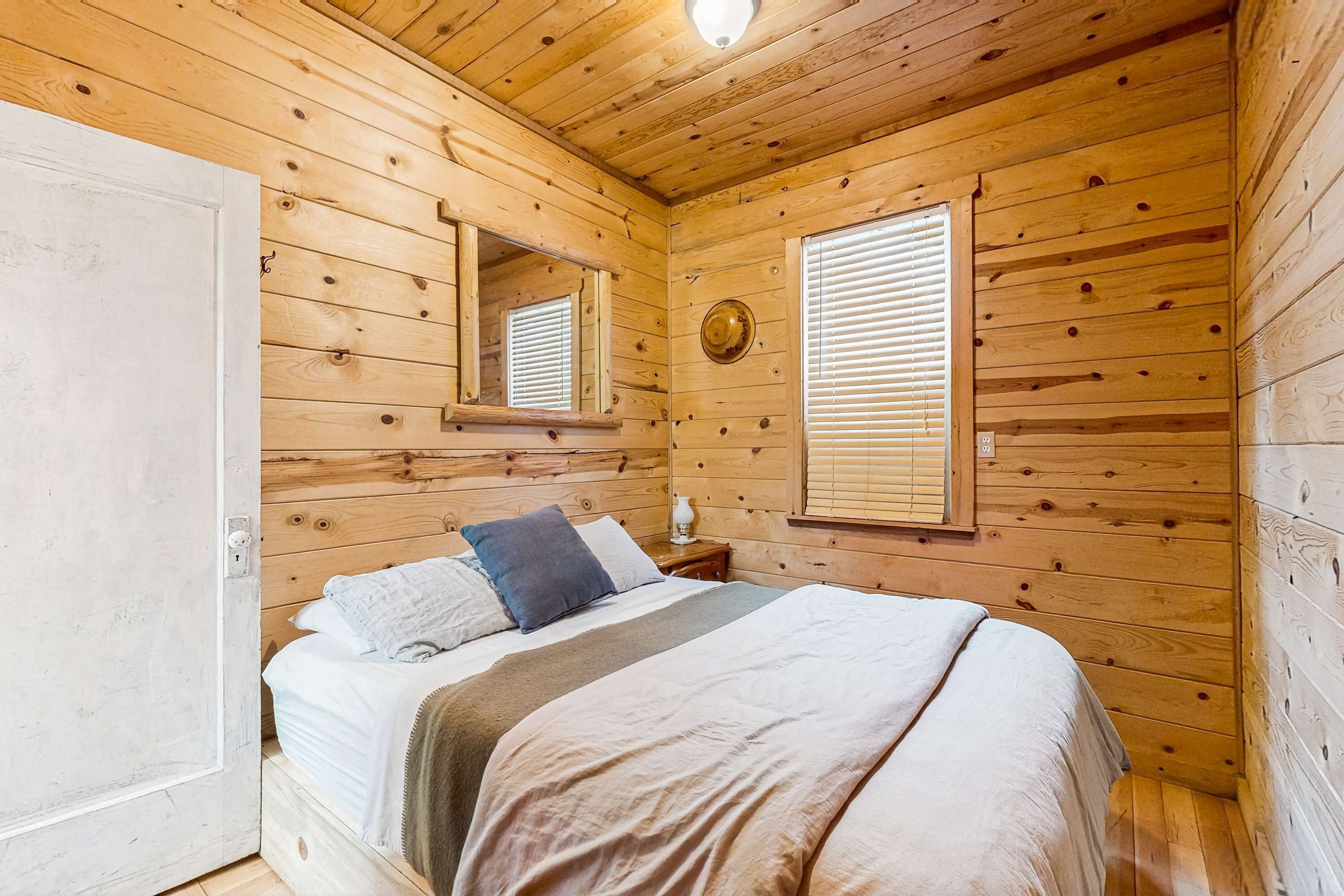 A wooden cabin bedroom with a bed, a nightstand, a mirror, and a window with blinds.