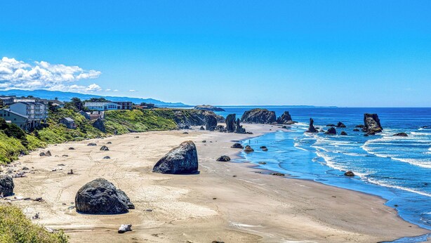 Une plage avec de gros rochers, une eau d’un bleu limpide et des maisons en arrière-plan.
