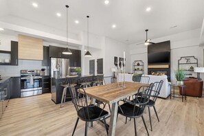 A modern kitchen with a central island, dining area with a wooden table and chairs, and a living area with a sofa and TV. - 2 Luxury King Suites + Hot Tub | Fire Pit • Pools (Hollister)