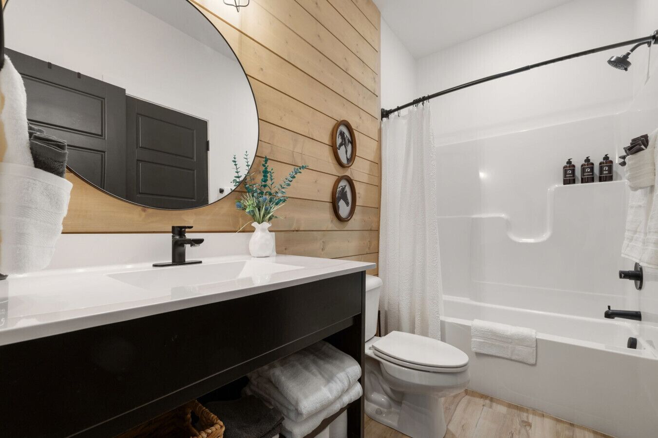 A modern bathroom with a wooden panel wall, a large mirror, a white sink, a black vanity, a toilet, and a glass-enclosed shower.