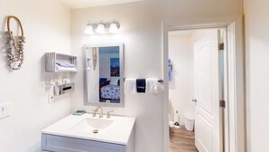 Un baño con lavamanos, espejo, toallero y una puerta que da a otra habitación.