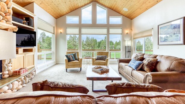 A spacious living room with a high ceiling, large windows, a stone wall, and leather furniture. - Exquisite home with hot tub, golf on-site, pool, tennis, & washer&dryer (Eagle Crest)