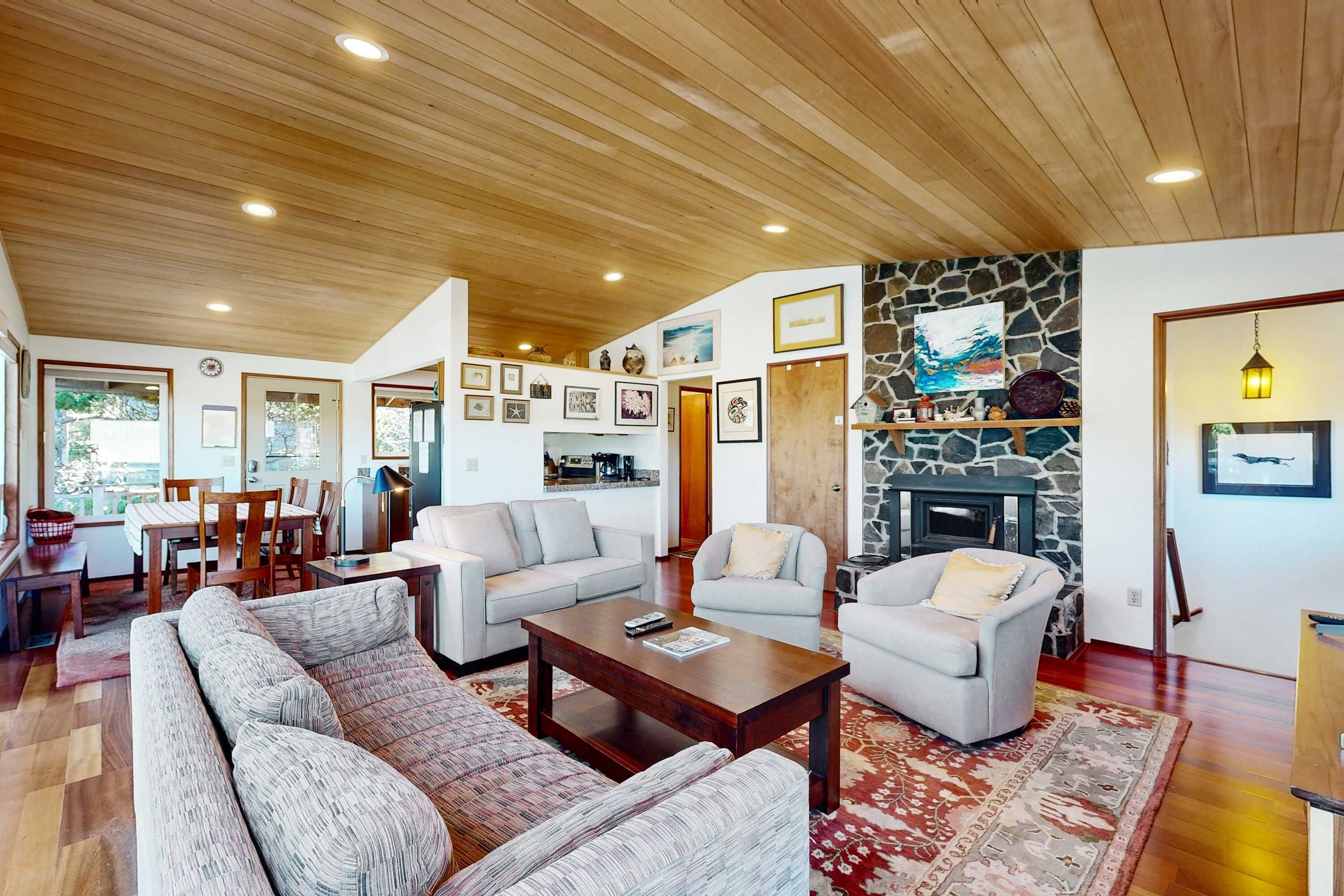 A spacious living room with a stone fireplace, wooden ceiling, and various framed pictures on the walls.