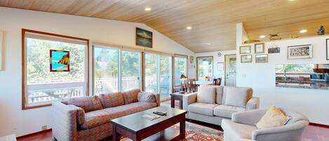A spacious living room with a wooden ceiling, large windows, a sofa, a coffee table, and a fireplace.