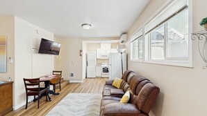 Una sala de estar con sofá, una mesa de comedor con sillas, un televisor montado en la pared y una cocina al fondo.