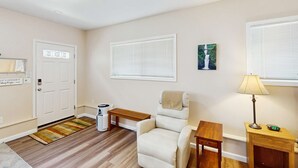 Una habitación con puerta, un banco, una silla, una mesita, una lámpara y una ventana con persianas.