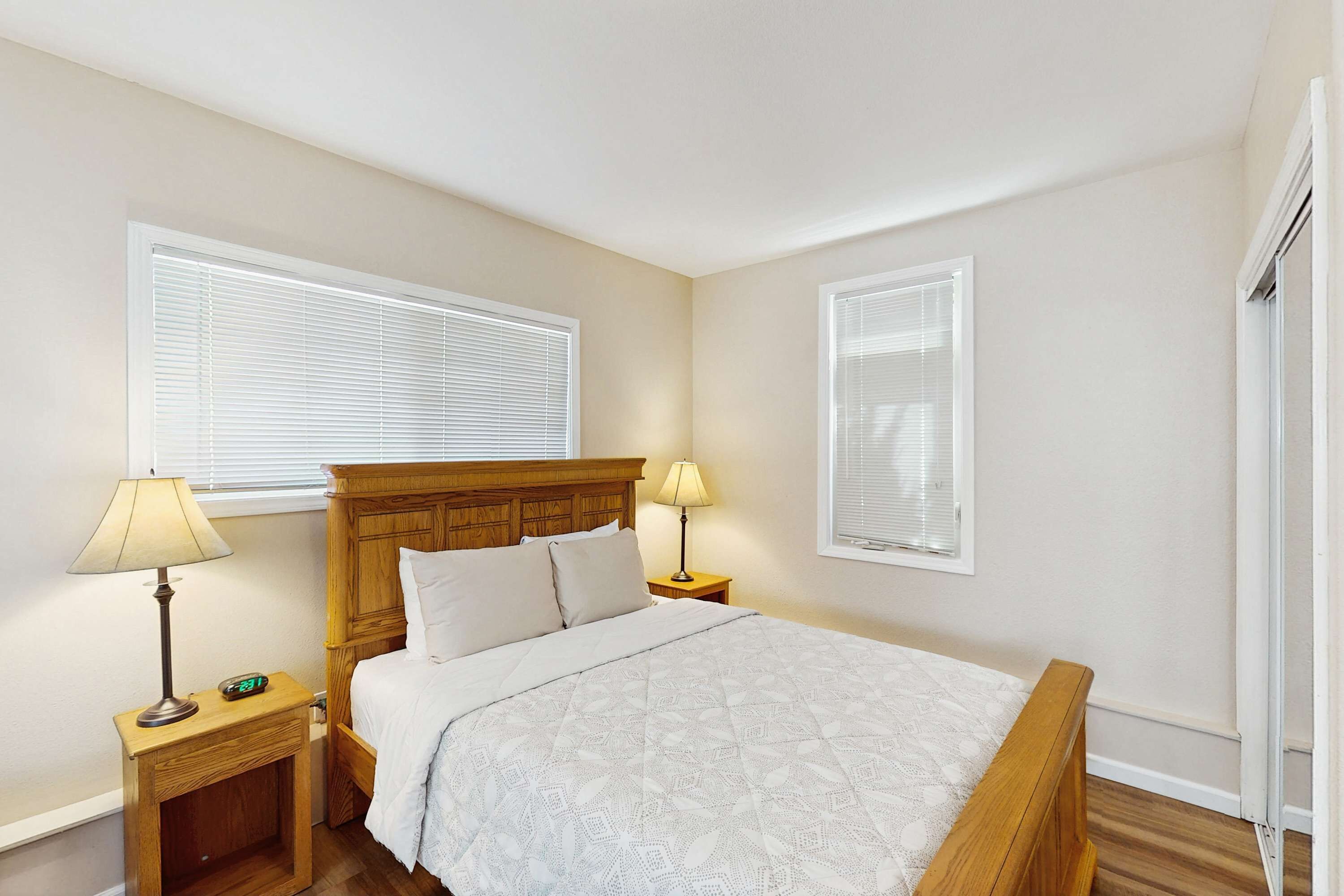 Un dormitorio con una cama de madera, dos mesitas de noche con lámparas y dos ventanas con persianas.