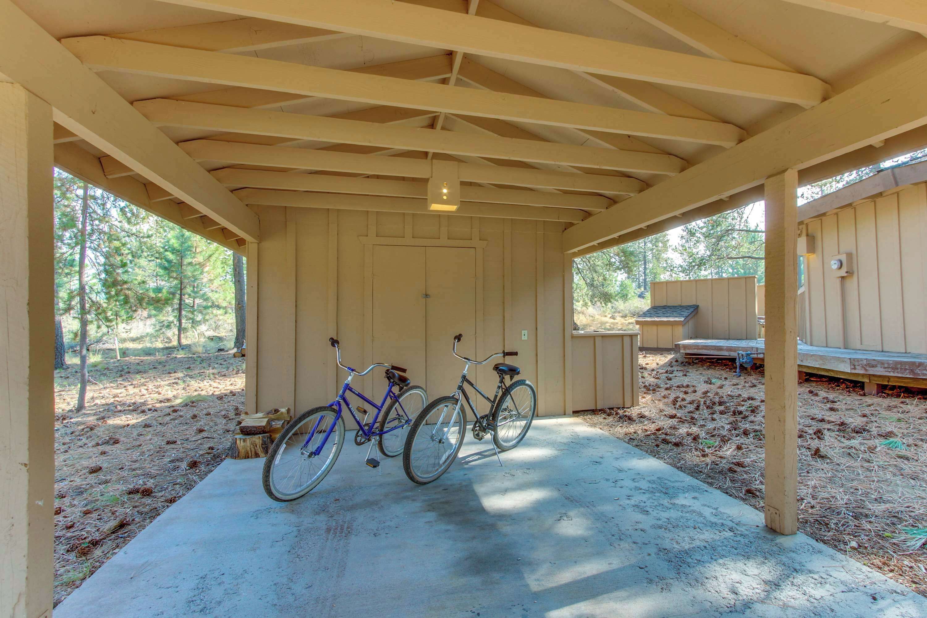 Un espace extérieur abrité où sont garés deux vélos sous une structure en bois. Le sol est en béton et, au fond, on aperçoit un bâtiment en bois.