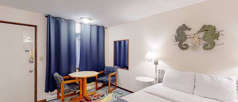 A hotel room with a bed, a desk with a chair, a small table, a lamp, and a decorative wall piece.